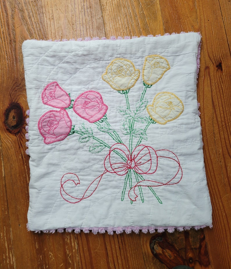 May include: A white pillow with a pink and yellow embroidered floral design. The pillow has a pink crocheted trim around the edges.