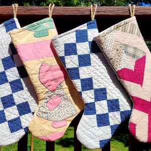 Puede incluir: Cinco calcetines de Navidad hechos a mano colgados de una barandilla de madera. Los calcetines presentan varios patrones acolchados, incluyendo cuadros azules y blancos, un corazón rosa y diseños a rayas. Cada calcetín está suspendido por un lazo de cuerda de yute.