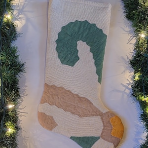 May include: A quilted Christmas stocking with a cream-colored background and patches of green, tan, and yellow. The stocking is decorated with a green wreath design. A jute loop is attached for hanging. The image is framed with a garland of green pine and white lights.
