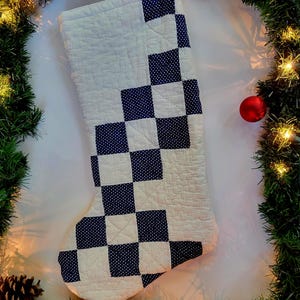 May include: A quilted Christmas stocking with a white background and a navy blue checkerboard pattern. The stocking is surrounded by a festive green garland with pinecones, a red ornament, and string lights.
