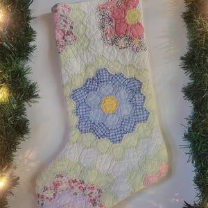 May include: A Christmas stocking crafted from a patchwork quilt featuring a hexagonal pattern. The design displays various floral and geometric fabric prints in soft shades of pink, blue, yellow, green, and white, stitched together in a flower motif.