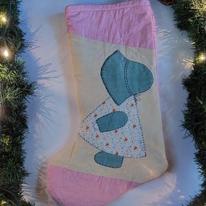May include: A handmade Christmas stocking with a quilted design. The stocking features a light pink top and bottom, with a cream-colored center panel. A blue and white applique of a figure in a dress is on the front. A jute loop is attached for hanging.