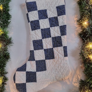 Puede incluir: Un calcetín navideño acolchado con un patrón de cuadros que alterna cuadrados blancos sólidos y cuadrados azul oscuro con un pequeño estampado de lunares blancos. La tela tiene una textura de cuadrícula cosida en toda la superficie.