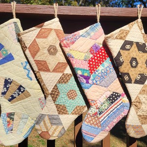 Op de afbeelding: Vier handgemaakte kerstsokken met unieke patchworkontwerpen. Elke sok heeft een ander kleurenschema en patroon, waaronder sterren en waaiervormen, met ophanglussen van touw.