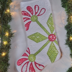 May include: White Christmas stocking with a quilted floral design in pink and green. The flowers have pink petals and green centers and stems. The stocking is decorated with a diamond pattern in green.