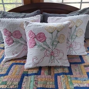 May include: Four white throw pillows with embroidered floral designs. The pillows have a ruffled edge and are arranged on a patchwork quilt.
