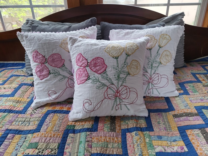 May include: Four white throw pillows with embroidered floral designs. The pillows have a ruffled edge and are arranged on a patchwork quilt.