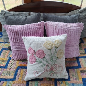 May include: A white cushion with embroidered pink and yellow flowers and a red bow. The cushion is on a bed with a patchwork quilt and several pink and grey cushions.