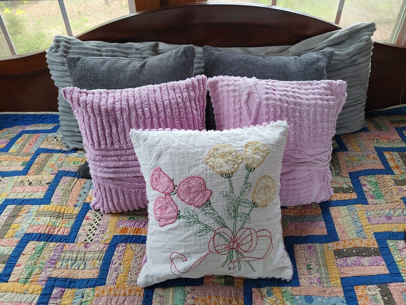 May include: A white cushion with embroidered pink and yellow flowers and a red bow. The cushion is on a bed with a patchwork quilt and several pink and grey cushions.