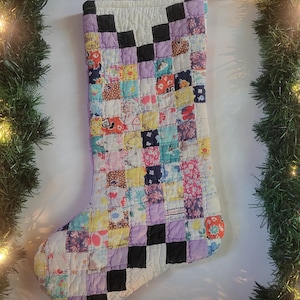 Puede incluir: Un calcetín navideño acolchado hecho de un mosaico de pequeños cuadrados de tela multicolor con varios patrones florales y geométricos, con un puño blanco y un diseño de cuadrícula que alterna cuadrados negros, lavanda y tonos pastel.