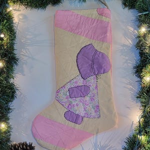 May include: A handmade Christmas stocking with a quilted design. The stocking features a light pink toe and heel, a beige body with a diamond pattern, and a purple applique of a figure with floral fabric accents. A loop is attached for hanging.