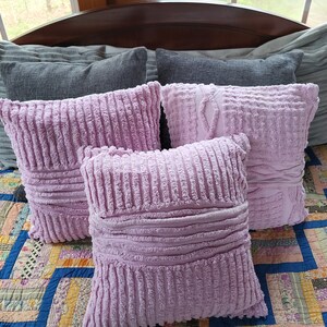 May include: Four decorative throw pillows on a bed. Two are grey and two are a soft, light purple. The purple pillows have a textured, ribbed fabric.