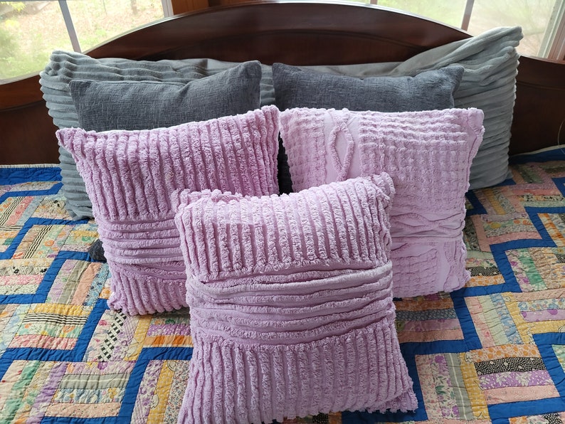 May include: Four decorative throw pillows on a bed. Two are grey and two are a soft, light purple. The purple pillows have a textured, ribbed fabric.