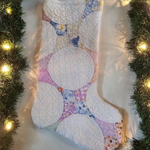 Puede incluir: Un calcetín navideño acolchado con fondo blanco y un patrón circular repetitivo compuesto por varios parches de tela con motivos florales y geométricos pequeños en tonos azul, rosa, naranja, morado y verde con un lazo para colgar.