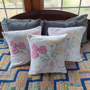May include: Four embroidered cushion covers with floral designs. Two cushions feature pink flowers and two cushions feature yellow flowers. The cushions are arranged on a patchwork quilt.