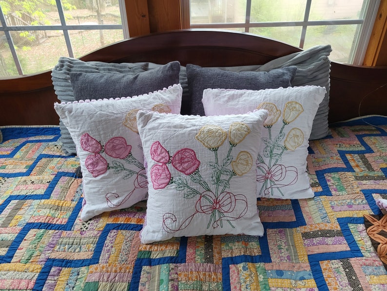 May include: Four embroidered cushion covers with floral designs. Two cushions feature pink flowers and two cushions feature yellow flowers. The cushions are arranged on a patchwork quilt.