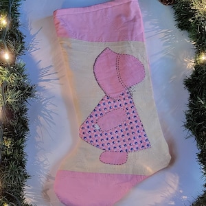 Puede incluir: Un calcetín de Navidad hecho a mano con una combinación de colores rosa y crema. El calcetín presenta una aplicación rosa de una niña con gorro y vestido con un estampado floral azul. El calcetín está adornado con tela rosa.