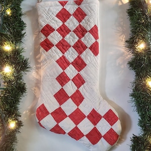 Puede incluir: Un calcetín navideño acolchado con una base de tela blanca y un patrón de diamantes rojos cosido en la parte delantera. El calcetín incluye un pequeño lazo de cuerda de yute de color natural en la esquina superior para poder colgarlo.
