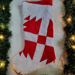 May include: A quilted Christmas stocking featuring a geometric pattern of alternating red and white fabric blocks. The stocking has a textured, stitched surface and includes a small, tan-coloured jute twine loop at the top for hanging purposes.