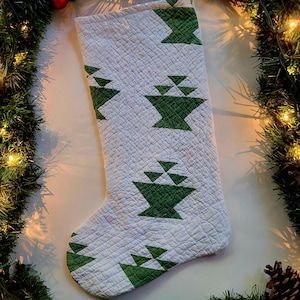 May include: White Christmas stocking with a green geometric tree pattern. The stocking is quilted and has a loop for hanging. The background includes a green garland with lights and a pinecone.