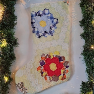 Puede incluir: Un calcetín navideño acolchado que presenta un mosaico de piezas de tela hexagonales en amarillo pálido, blanco y verde claro. El calcetín muestra dos motivos florales destacados, uno en cuadros vichy azules y otro en rojo, con centros amarillos.