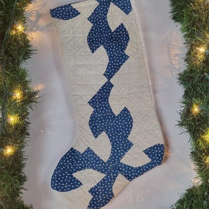May include: A quilted Christmas stocking with a cream-colored background and blue star-patterned accents. The stocking is decorated with a geometric design and has a jute loop for hanging. The overall theme is holiday decor.