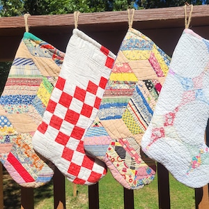 Op de afbeelding: Vier gewatteerde kerstsokken hangen op een rij. De eerste en derde sok hebben een veelkleurig patchworkontwerp met diverse stofpatronen. De tweede sok heeft een rood-wit schaakbordpatroon. De vierde sok is wit van kleur.