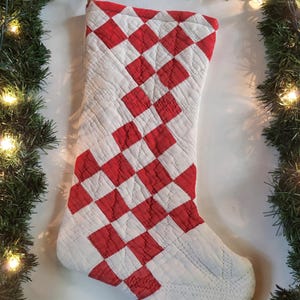 Puede incluir: Un calcetín navideño acolchado con un patrón de cuadros en forma de diamante rojo y blanco. El calcetín está confeccionado con costuras visibles en toda la tela e incluye un sencillo lazo de cuerda de yute en la parte superior para colgarlo.