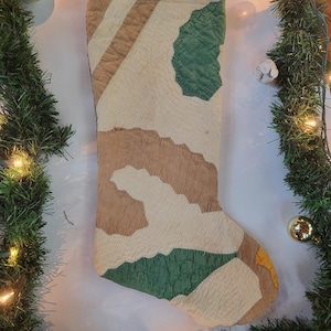 May include: A Christmas stocking with a patchwork design in shades of beige, brown, and green. The stocking has a quilted texture and a classic Christmas shape. The background includes festive greenery and lights.