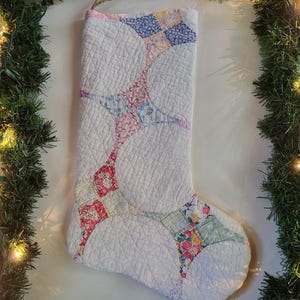 Könnte beinhalten: Ein gesteppter Weihnachtsstrumpf mit weißem Hintergrund und einem sich wiederholenden Muster aus ineinandergreifenden Kreisen und Quadraten in verschiedenen Blumenmustern in Blau, Rosa, Rot, Gelb und Grün, versehen mit einer einfachen Aufhängeschlaufe aus Garn.