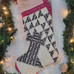 May include: Handmade Christmas stocking with a patchwork design. The stocking features a cream-colored background with black and gray triangles and patterned sections. It has a pink border and a loop for hanging.