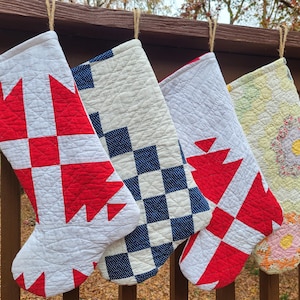 Puede incluir: Cuatro calcetines navideños acolchados con diferentes patrones, colgados de una barandilla de madera. Los diseños incluyen diseños geométricos rojos y blancos, cuadros azules y blancos y motivos florales. Cada calcetín está suspendido por un lazo de cuerda.