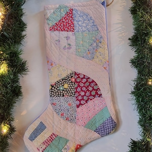 Könnte beinhalten: Ein gesteppter Weihnachtsstrumpf aus verschiedenen Stoffstücken in den Farben Rosa, Blau, Rot, Gelb und Schwarz. Der Strumpf weist ein geschwungenes Patchwork-Design mit sichtbaren Nähten und eine kleine Schlaufe aus Jute zum Aufhängen auf.
