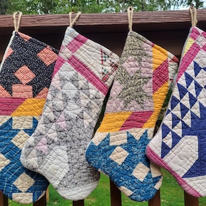 Può includere: Quattro calze natalizie trapuntate fatte a mano con vari motivi e colori. Ogni calza ha un design unico, tra cui forme geometriche, stelle e motivi floreali. Le calze sono realizzate in tessuto nei toni del blu, rosso, giallo e bianco e sono appese a una ringhiera di legno.