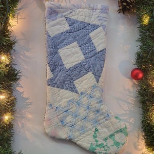 Puede incluir: Un calcetín navideño acolchado con un diseño de patchwork en tonos azules, blancos y verdes. El calcetín presenta varios patrones geométricos y está adornado con tela azul claro. El fondo incluye una guirnalda festiva y una bola roja.