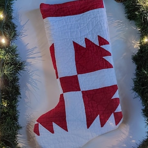 May include: A quilted Christmas stocking in red and white, featuring a geometric pattern. The stocking is decorated with a red and white design. The background includes a green garland with lights and a pinecone.
