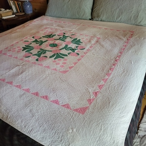 以下が含まれることがあります： 中央にピンクと緑の花柄が施された白いキルト。キルトは木製のヘッドボードと2つの薄い青色の枕のあるベッドの上に置かれています。