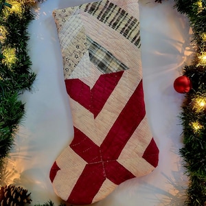 May include: A handmade Christmas stocking with a patchwork design. The stocking features a red and cream color scheme with a geometric pattern. The top of the stocking has a loop for hanging. The background includes a Christmas garland and lights.