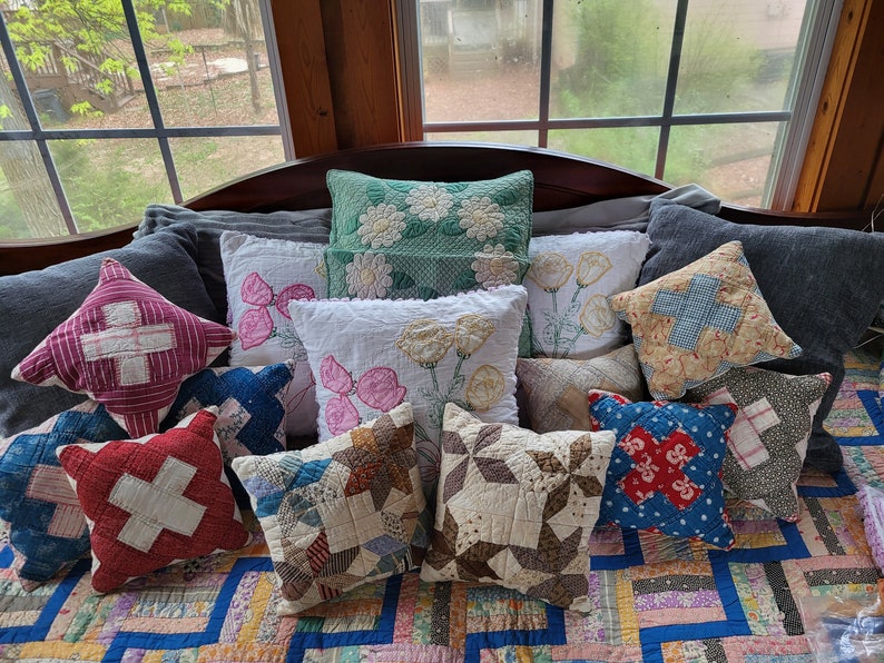 May include: A collection of vintage patchwork cushions in various colours and patterns, including red, blue, green, and white. The cushions are arranged on a bed with a patchwork quilt.