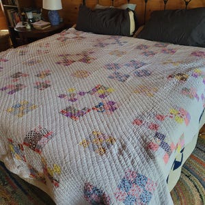 May include: A vintage quilt with a white background and colorful patchwork designs. The quilt features a repeating pattern of floral and geometric shapes in shades of pink, purple, yellow, and blue. The quilt is on a bed with dark gray pillows.