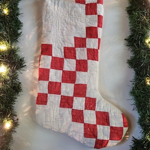Peut inclure: Une chaussette de Noël matelassée avec un motif à carreaux rouges et blancs. Le tissu présente des coutures visibles sur toute la surface, créant un aspect texturé. Une petite boucle est fixée au coin supérieur pour la suspendre.