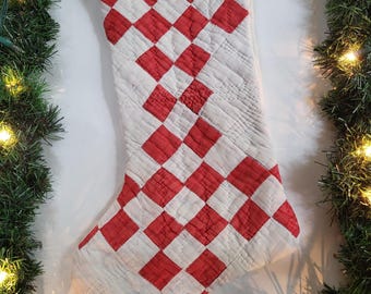 May include: A quilted Christmas stocking featuring a repeating diamond pattern in alternating red and white fabric squares. The stocking includes a small jute twine loop at the top for hanging, with visible stitching throughout the quilted surface.