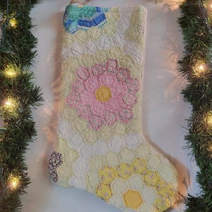 May include: A Christmas stocking crafted from quilted fabric featuring a patchwork of hexagonal shapes in pastel yellow, white, pink, and blue floral patterns. The stocking includes a small jute twine loop at the top for hanging purposes.