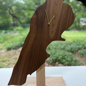 May include: A wooden clock shaped like a country's outline, featuring a dark brown wood grain. The clock has gold-colored hands and is mounted on a light-colored wooden base. The clock is approximately 18 inches tall.