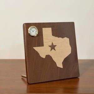 May include: A wooden desk clock featuring the state of Texas with a star in the center. The clock has a light wood Texas cutout on a dark wood background. A small, round clock with Roman numerals is mounted on the top left.