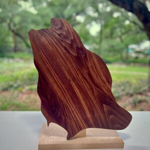 May include: A handcrafted wooden sculpture with a dark brown finish, mounted on a light-colored wooden base. The abstract form has a flowing, organic shape, showcasing the natural wood grain. The sculpture is displayed against a blurred outdoor background.