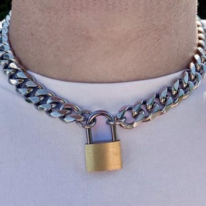 May include: A silver-colored chain necklace with large, interlocking links. The necklace features a gold-colored padlock clasp. The necklace is worn around the neck, against a white shirt.