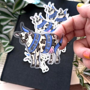 Op de afbeelding: Een transparante sticker met een cartoon-koe die een blauw lint draagt ​​met de tekst "I gotta go juju" en een ander blauw lint met de tekst "We got cows!" De koe is zwart-wit met een roze uier.
