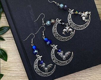 Könnte beinhalten: Drei Paar silberne Halbmond-Ohrringe mit baumelnden Fledermäusen und Kristallperlen. Die Ohrringe haben rote, blaue und grüne Kristalle.