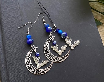 Puede incluir: Un par de pendientes de plata con un diseño de luna creciente y un dije de murciélago. Los pendientes están adornados con cuentas de vidrio azul.